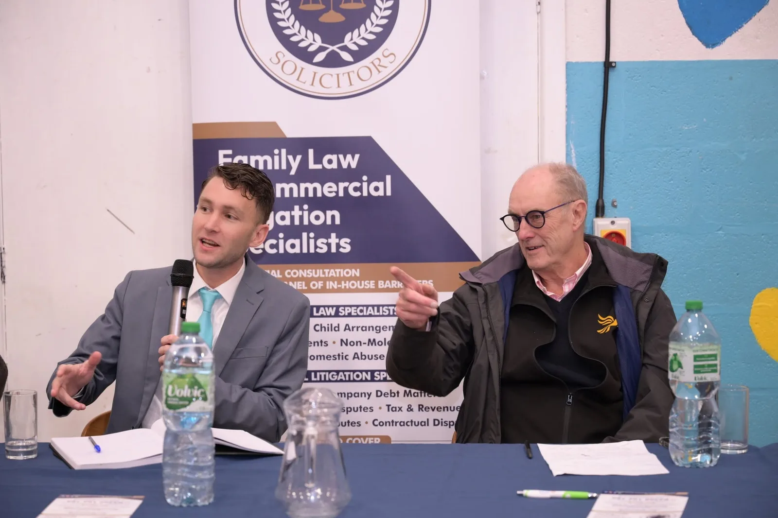 Two panellists at the table, one speaking into a microphone while the other gestures towards the room.